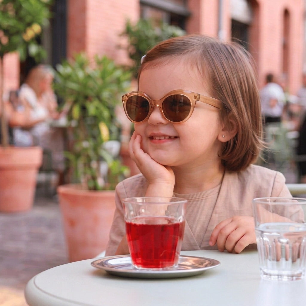 Petite fille à la terrasse d'un café portant des lunettes de soleil pour enfant forme papillon en acétate translucide couleur champagne avec verres marron.