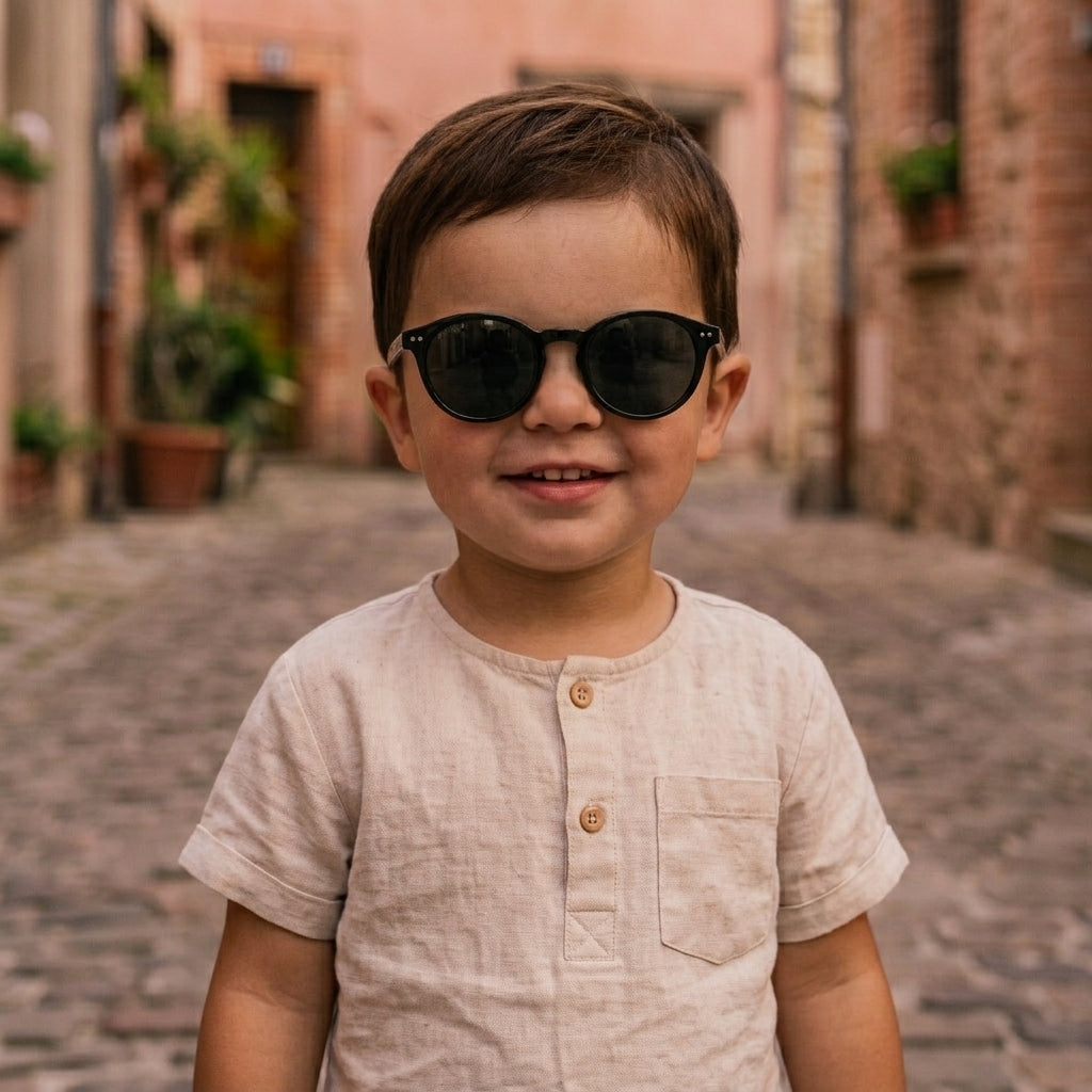 Petit garçon portant des lunettes de soleil enfant rondes noires avec verres foncés, vêtue d'un t-shirt en lin beige.