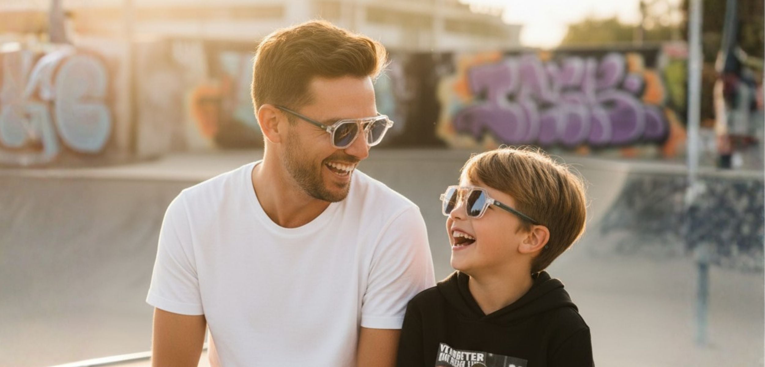 Portrait complice et solaire d'un père et de son fils partageant un éclat de rire dans un skatepark au coucher du soleil, portant tous les deux les lunettes assorties Kanumera C1 de Lumeya Suntribe au design transparent et moderne.