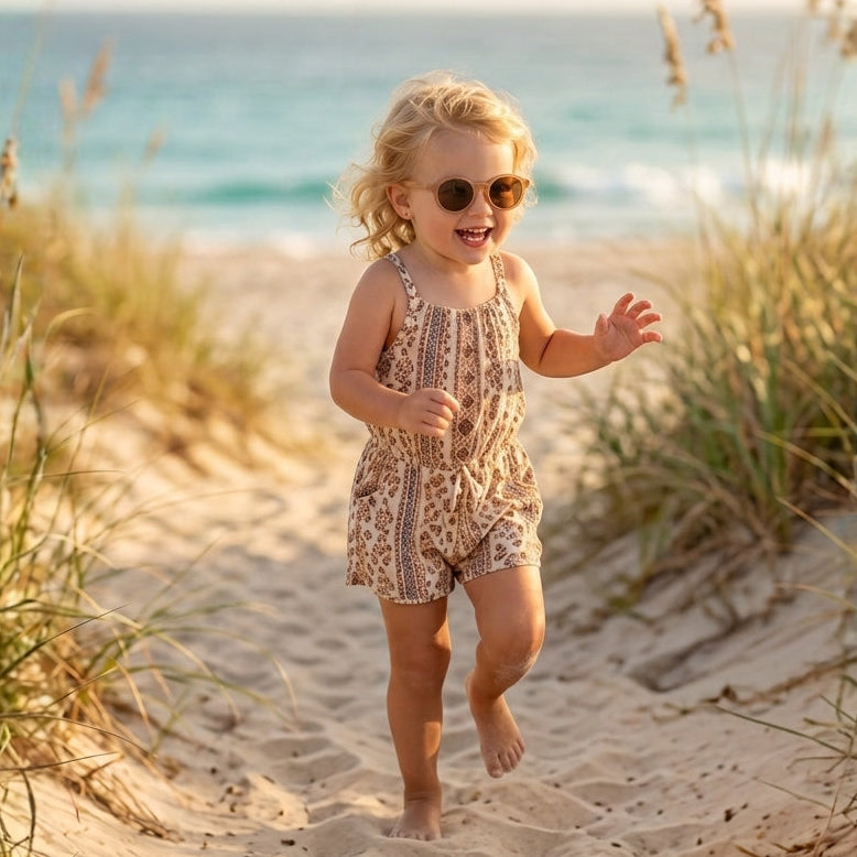 Petite fille sur la plage portant des lunettes de soleil rondes beiges et un combishort à motifs bohèmes.