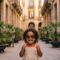Petite fille de 2 ans souriante dans une rue pavée, portant des lunettes de soleil ovales écaille de tortue à verres gris de catégorie 3 et une robe beige.