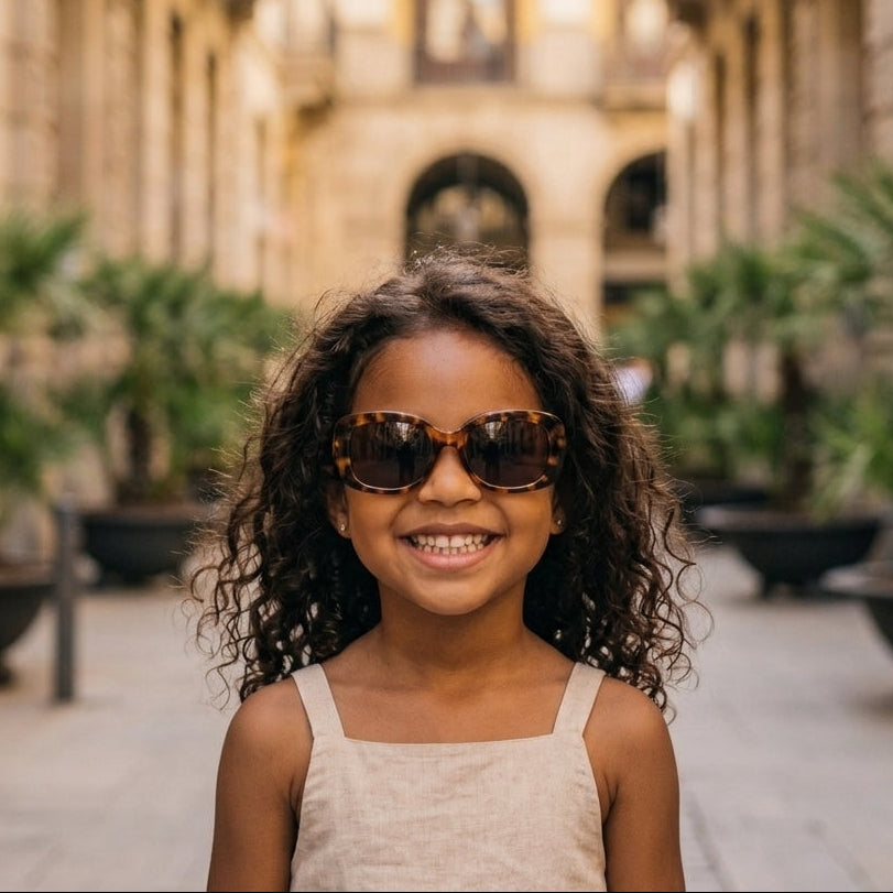 Portrait de face d'une petite fille métisse souriante, portant des lunettes de soleil à monture écaille de tortue et une tenue en lin beige, marchant dans une rue de ville ensoleillée.
